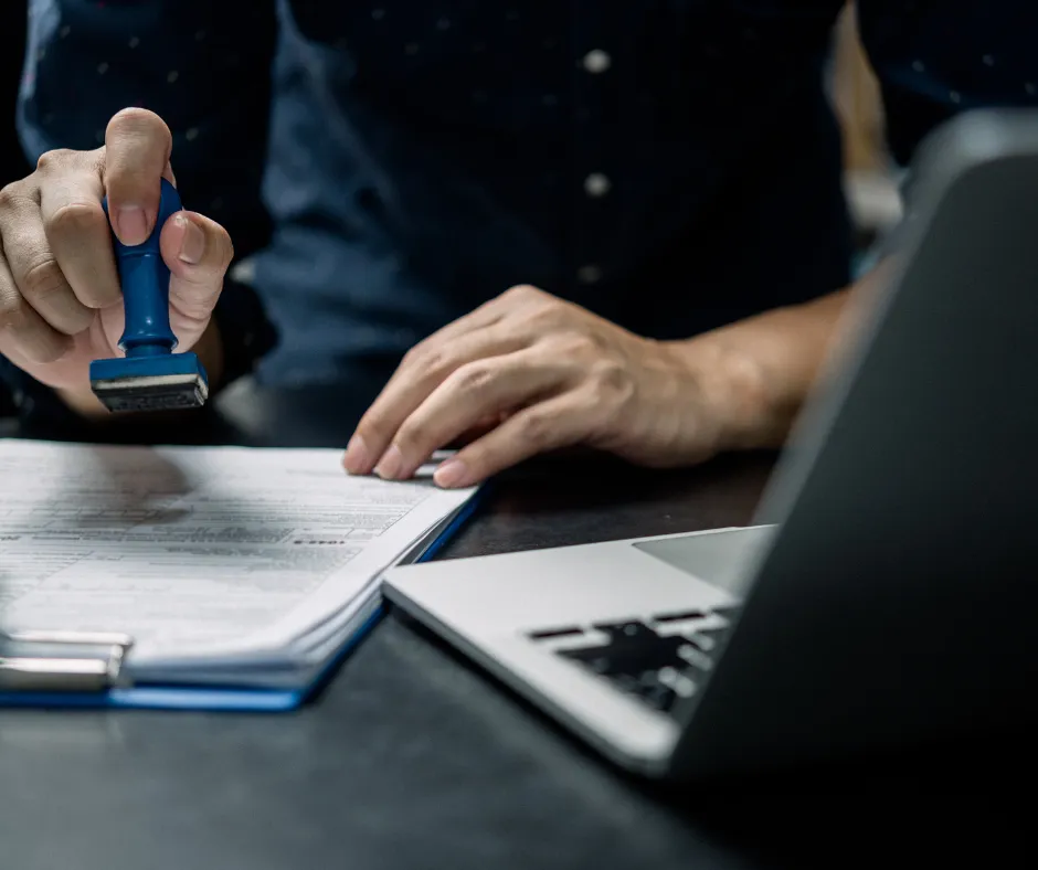 Person stamping document with blue stamp, laptop nearby.