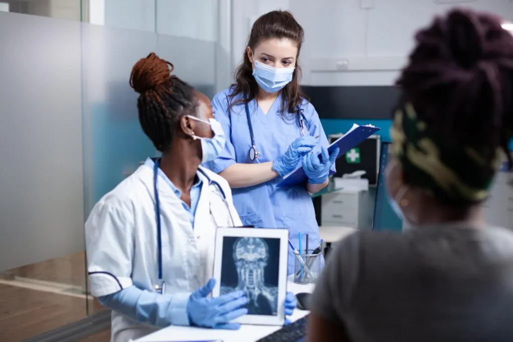 Three doctors in masks review medical imaging, discussing a patient's case.