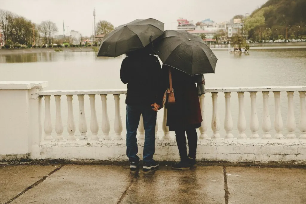 Couple holding hands under umbrellas by a lake.