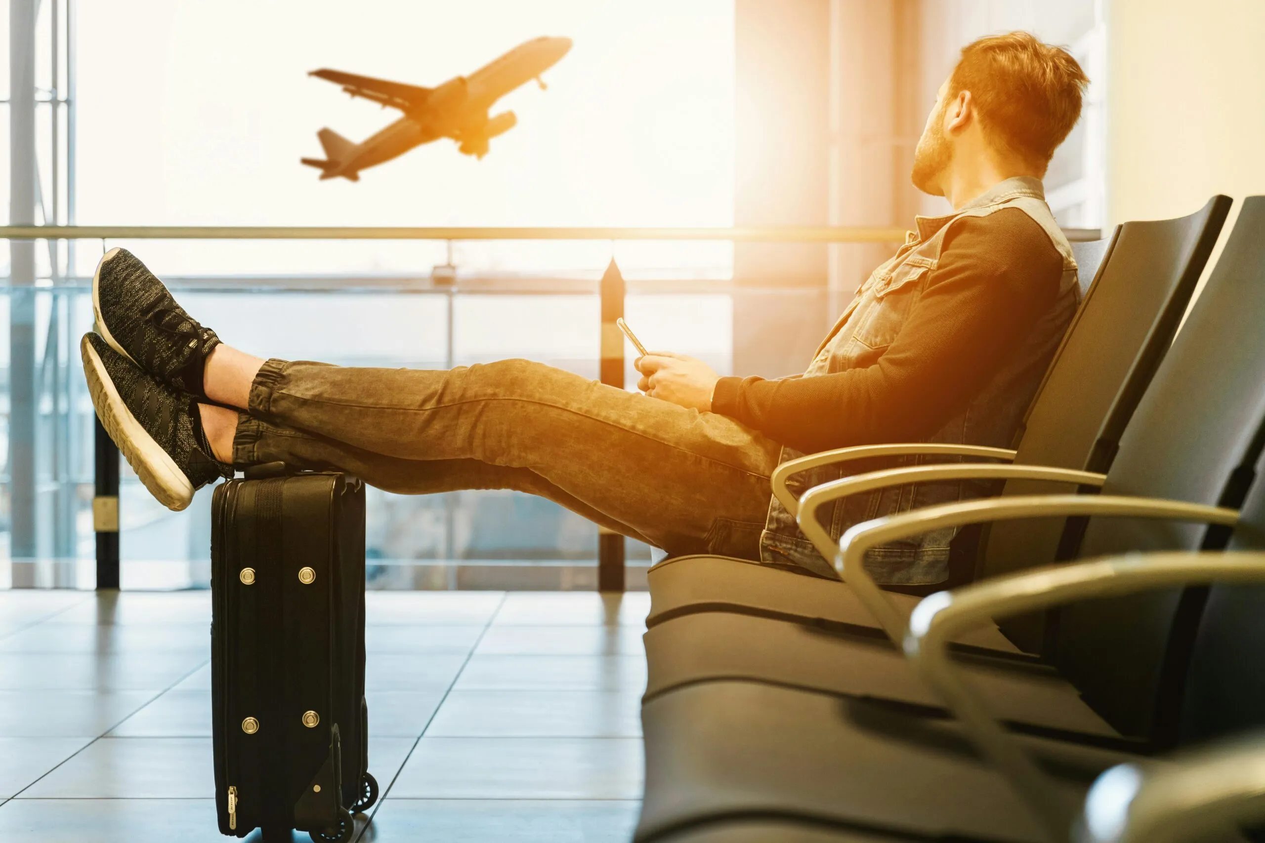 Man at airport with feet on luggage, watching a plane take off. Travel concept.