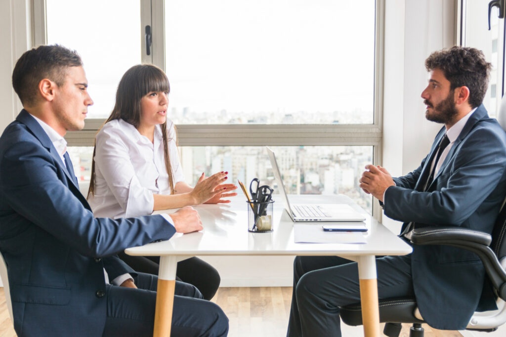 Three businesspeople in suits engaged in commercial mediation, seated around a table with a laptop.