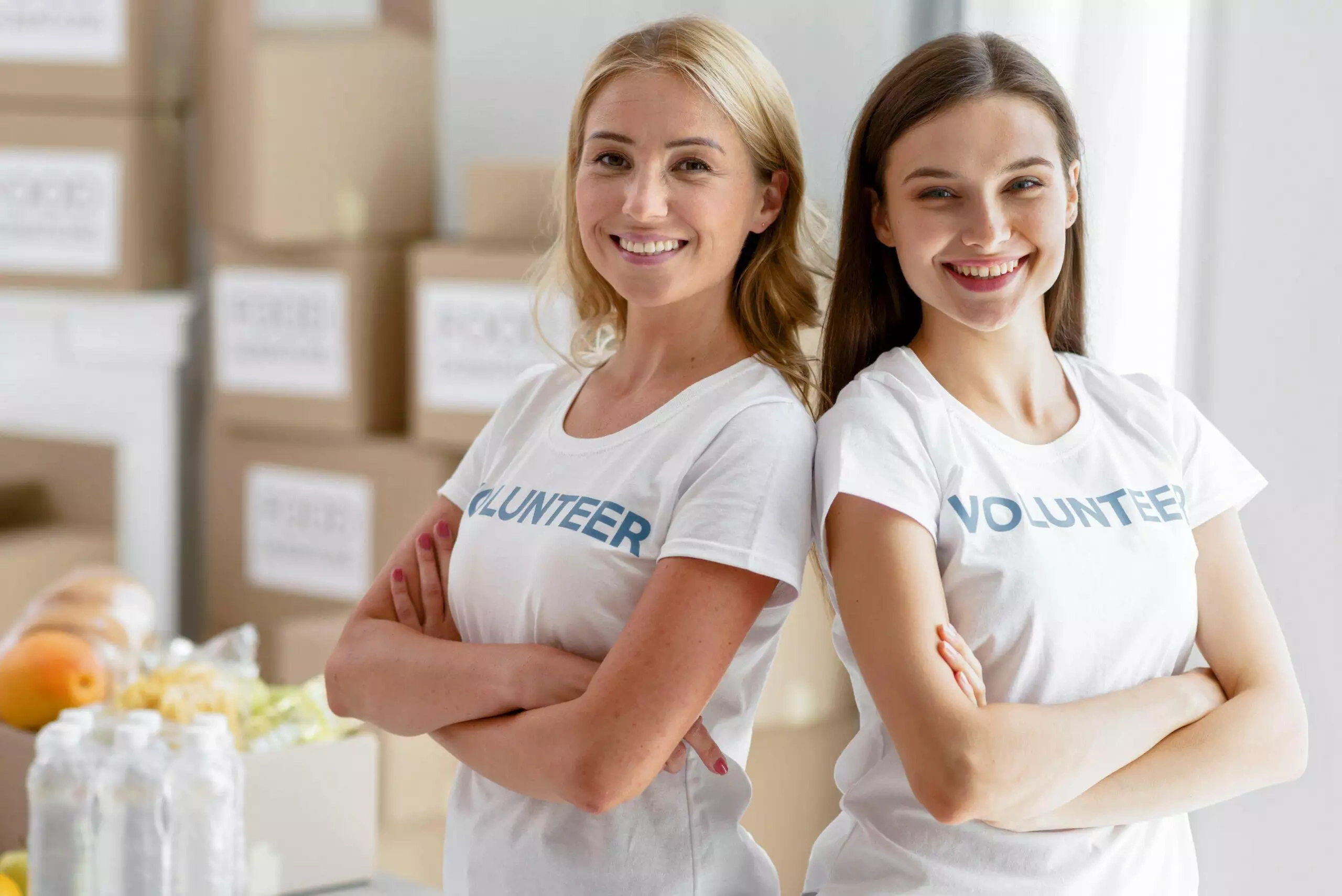 Two smiling female volunteers stand with arms crossed, wearing white 