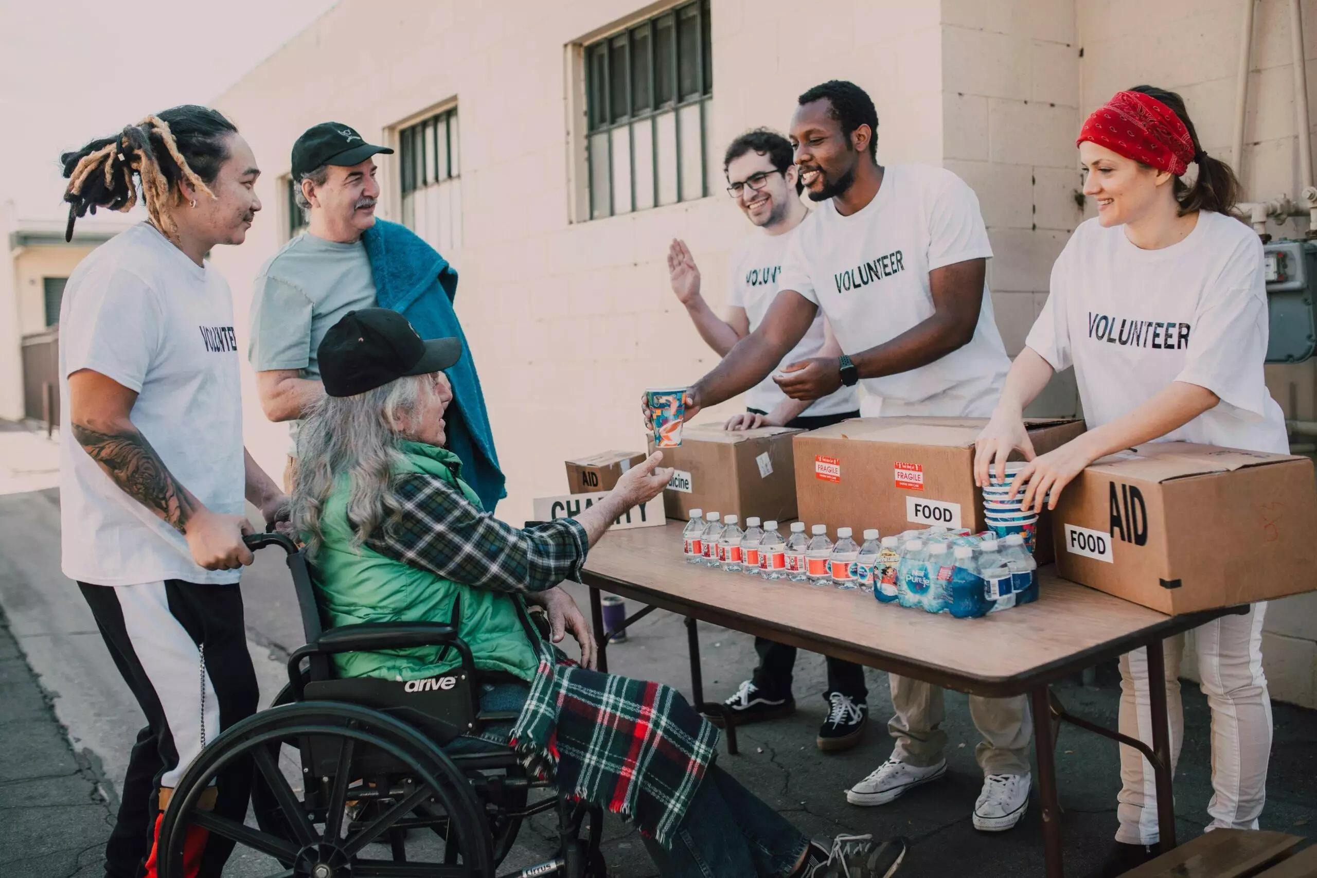 Volunteers distribute food and water to a person in a wheelchair. Boxes labeled