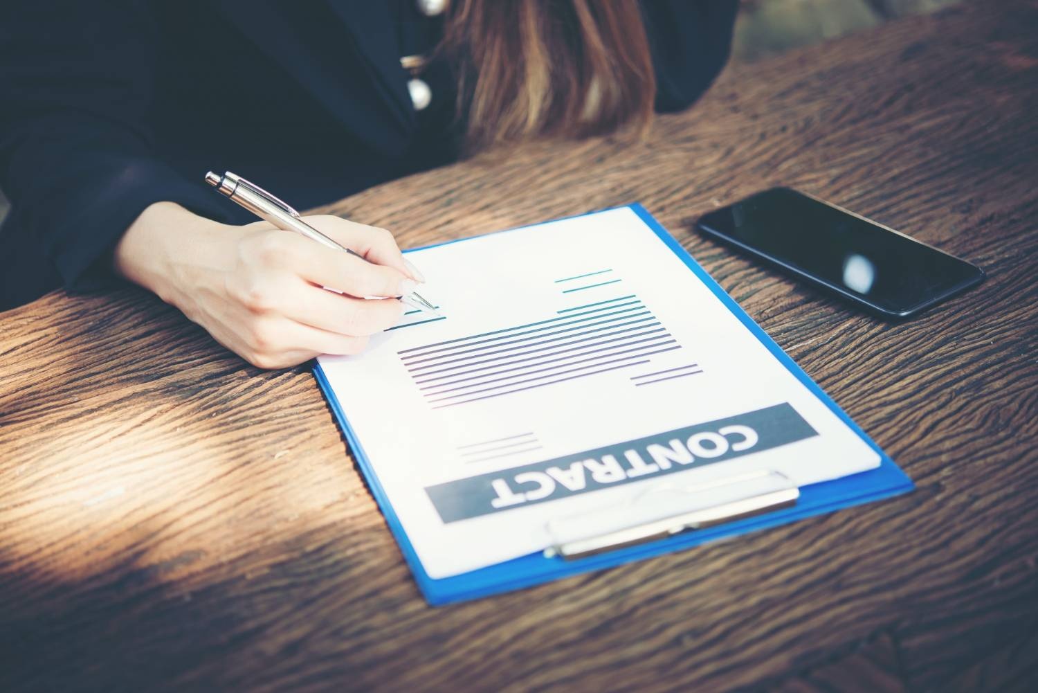 Signing a contract on a clipboard. Legal document being reviewed at a wooden desk, along with a phone.