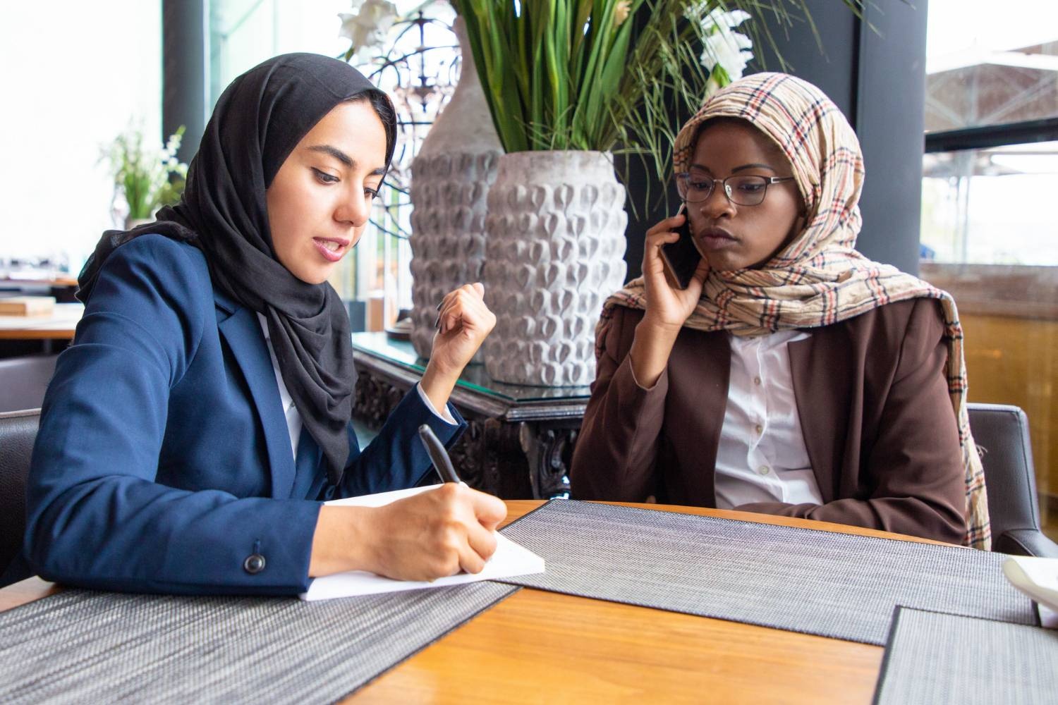 Two Muslim women discussing Islamic wills and estate planning, one taking notes, another on a call.