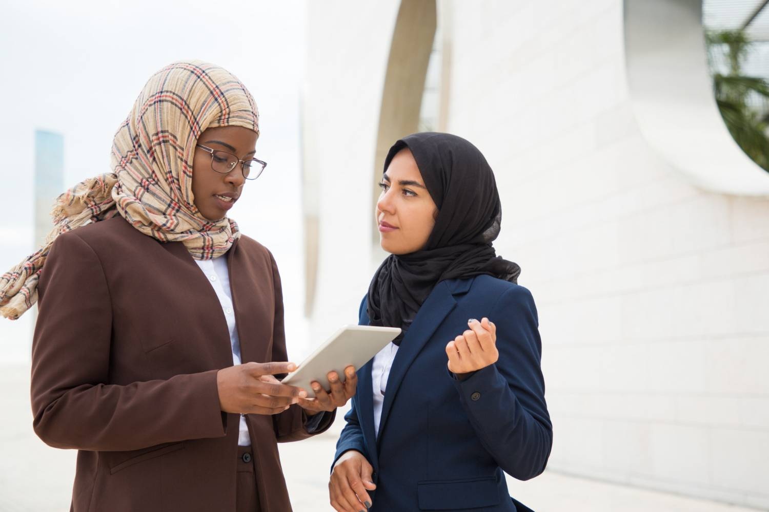 Two Muslim women in business attire review information on a tablet outdoors.
