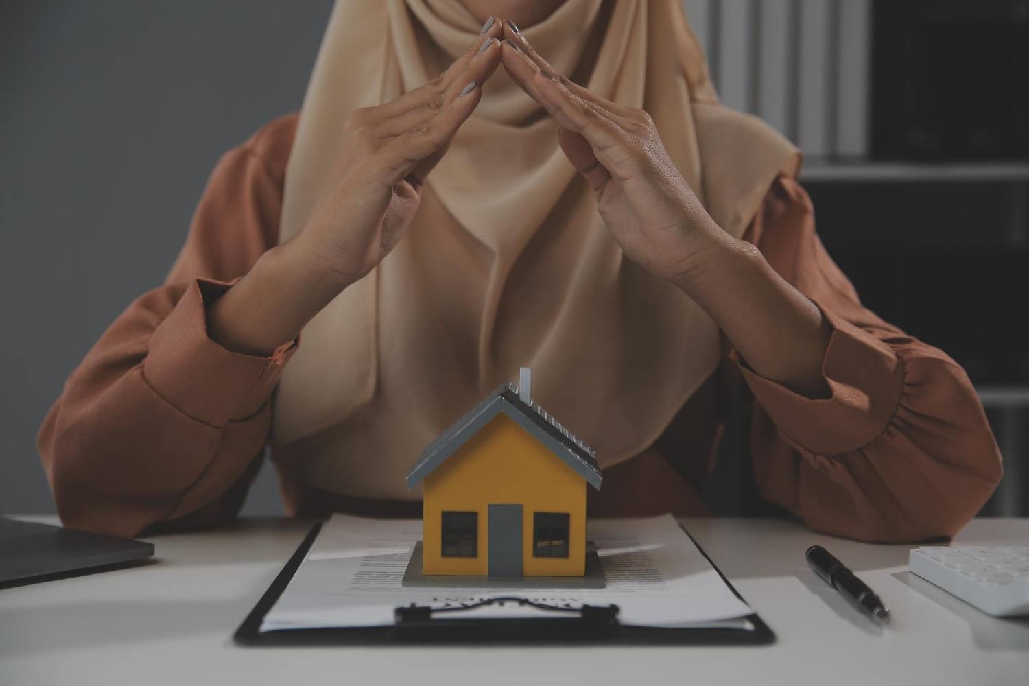 Islamic estate planning: Woman in hijab protects miniature house with hands.