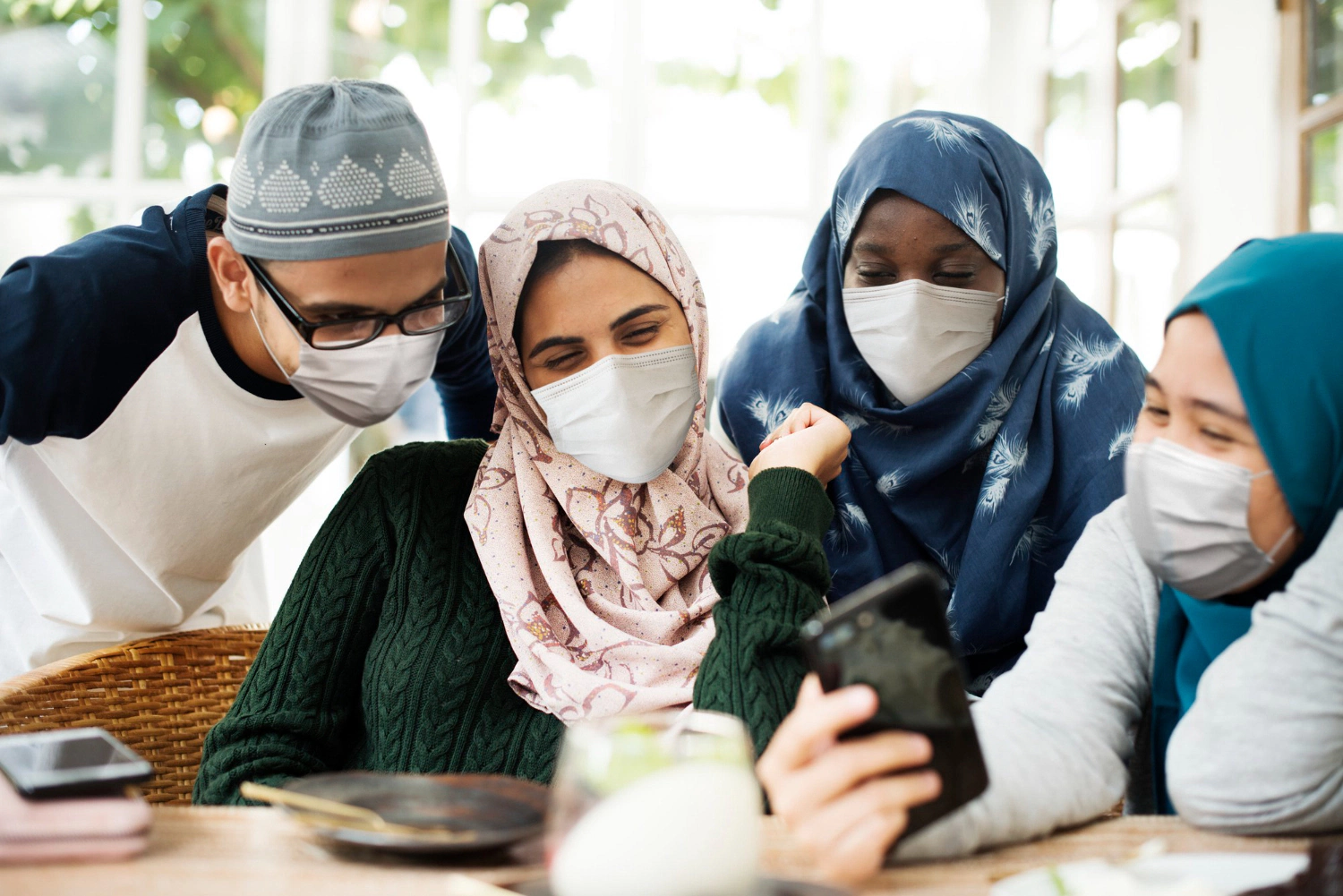 Diverse Muslim friends wearing masks look at phone together.